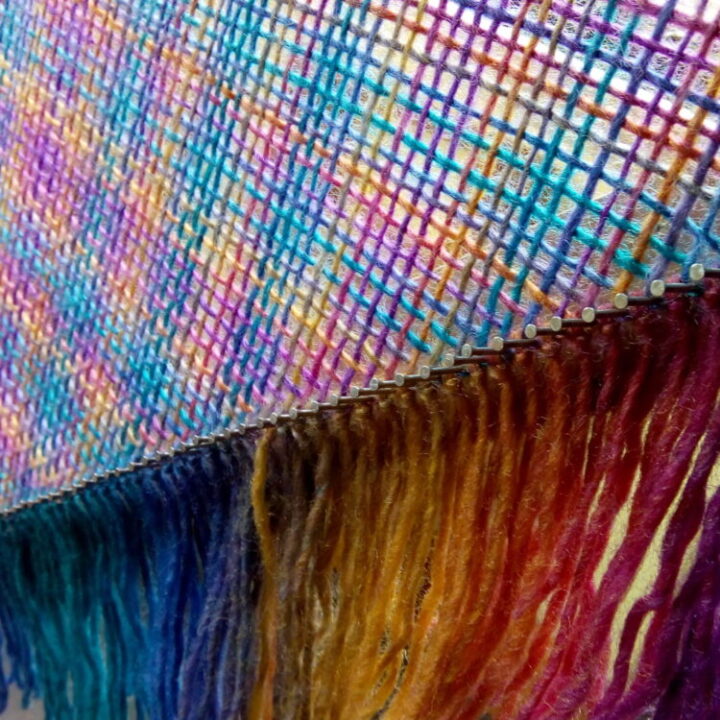Close up view of hand woven shawl with fringe in pinks, golds and blues before being taken off the triloom.