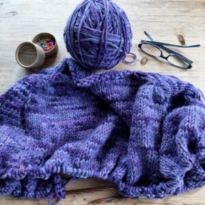 A ball of hand dyed yarn, a piece of knitting in progress with a pot of Cocoknits coloured stitch markers and a pair of glasses.