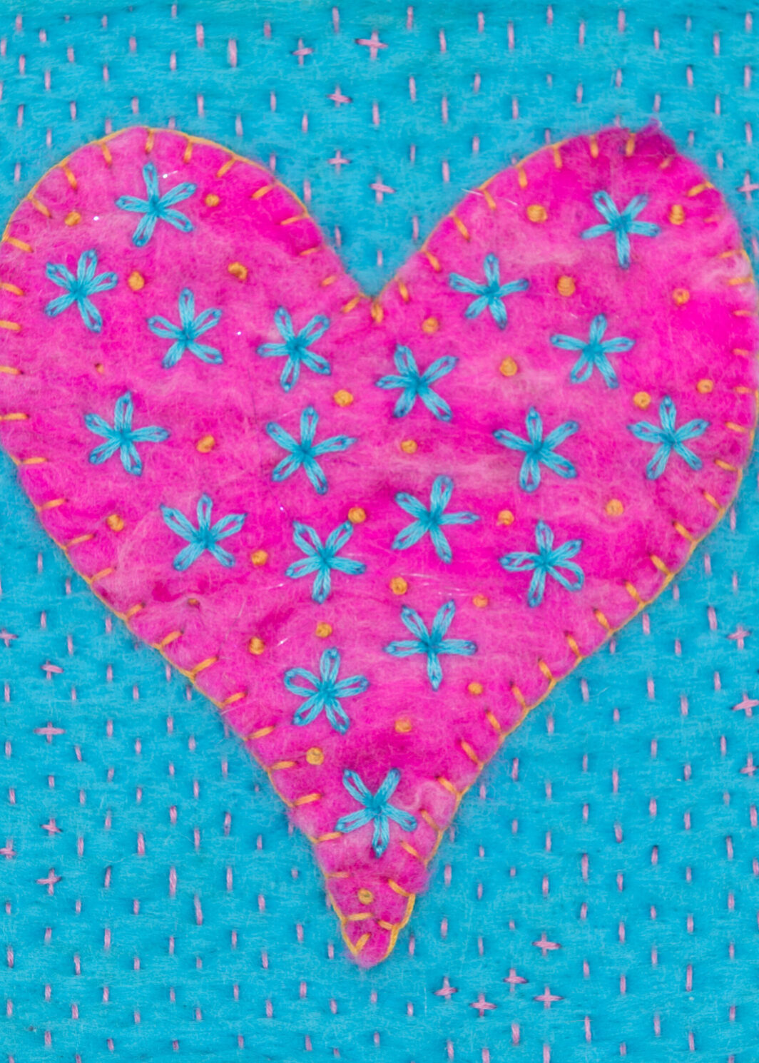 Greetings card image of a bright pink heart blanket stitched with golden yellow thread onto a bright turquoise felt background. The heart is embroidered with turquoise flowers and golden yellow French knots. The background is embroidered with tiny pink running stitch.