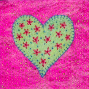 Greetings card image of a pale green heart blanket stitched with blue thread onto a bright pink background. The heart is embroidered with pink flowers and blue French knots. The background is embroidered with tiny pale green running stitch.