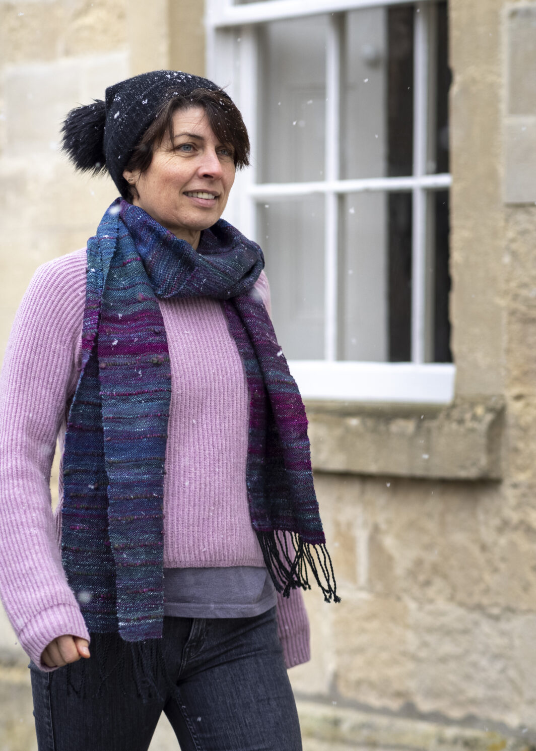 Woman with dark hair and black bobble hat is walking down the street past a window. She is wearing a lilac jumper and is modelling a hand woven silk scarf in oil slick colours of teal, blues and deep pink.