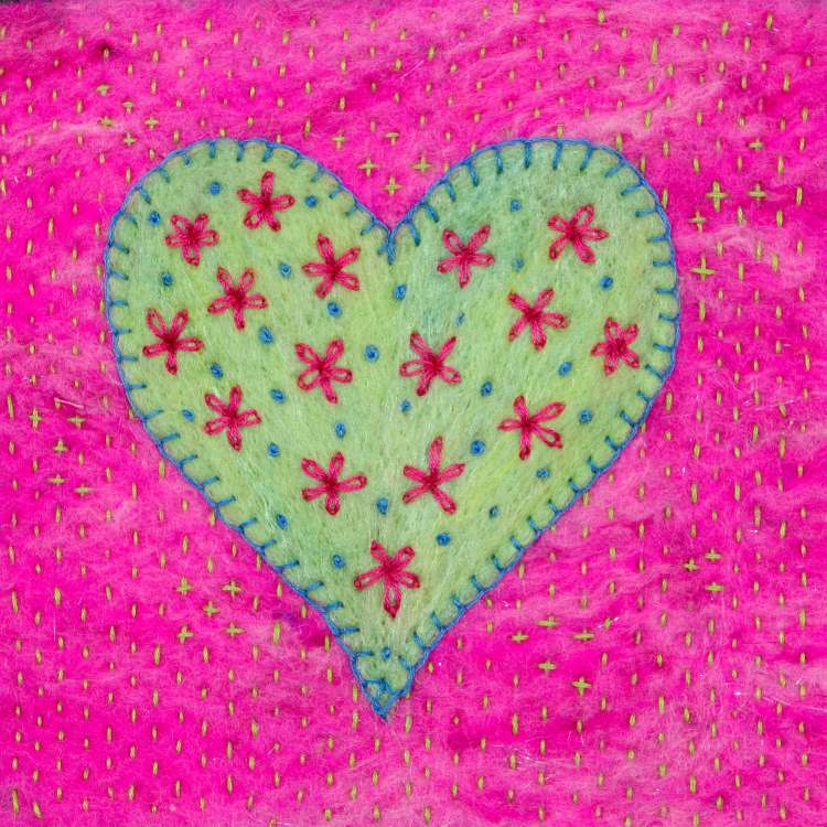 Greeting card image of a pale green heart blanket stitched with blue thread onto a bright pink background. The heart is embroidered with pink flowers and blue French knots. The background is embroidered with tiny pale green running stitch.