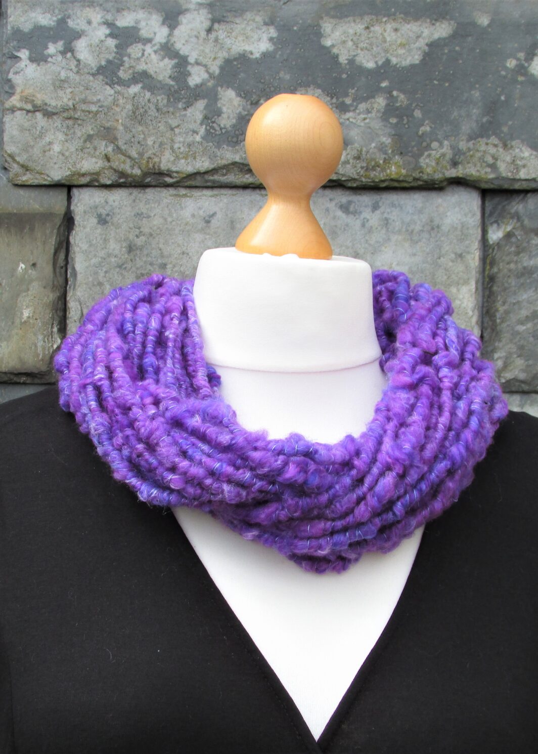 Tailor's dummy is modelling a bluey purple hand spun yarn cowl in front of a slate hung wall.
