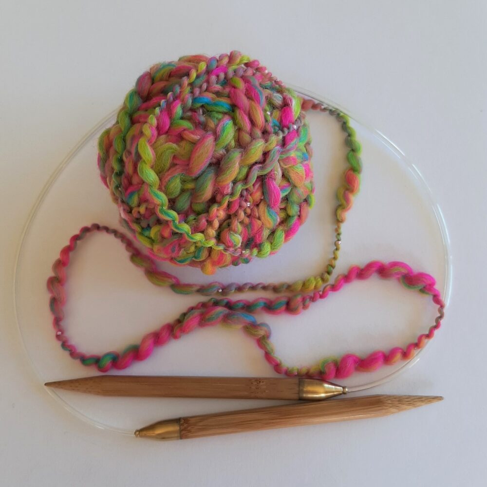 Ball of bright pink, yellow, turquoise and lime green thick and thin handspun yarn incorporation tiny pink beads and a pair of 10mm bamboo circular needles. Button to buy handspun beaded yarn.