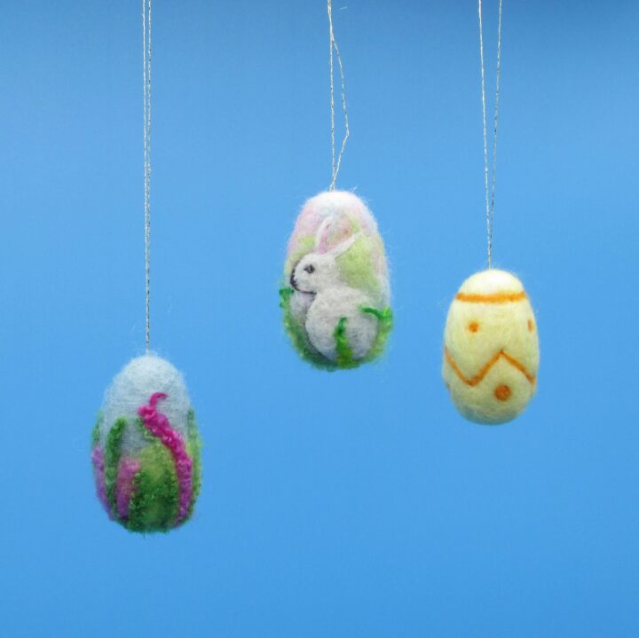 Three decorated needle felt easter eggs are suspended from golden thread in front of a sky blue background. the one on the left has a flower-like decoration, the middle egg has a rabbit and the one on the right is yellow with an orange abstract motif.