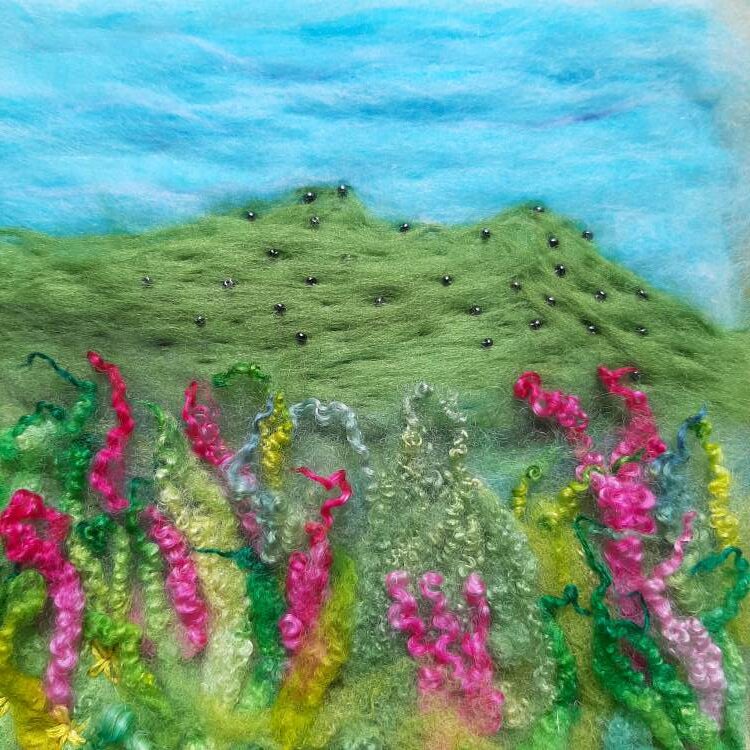 A needle felt image of blue sky, green Roughtor with foxgloves made of hand dyed locks in the foreground.