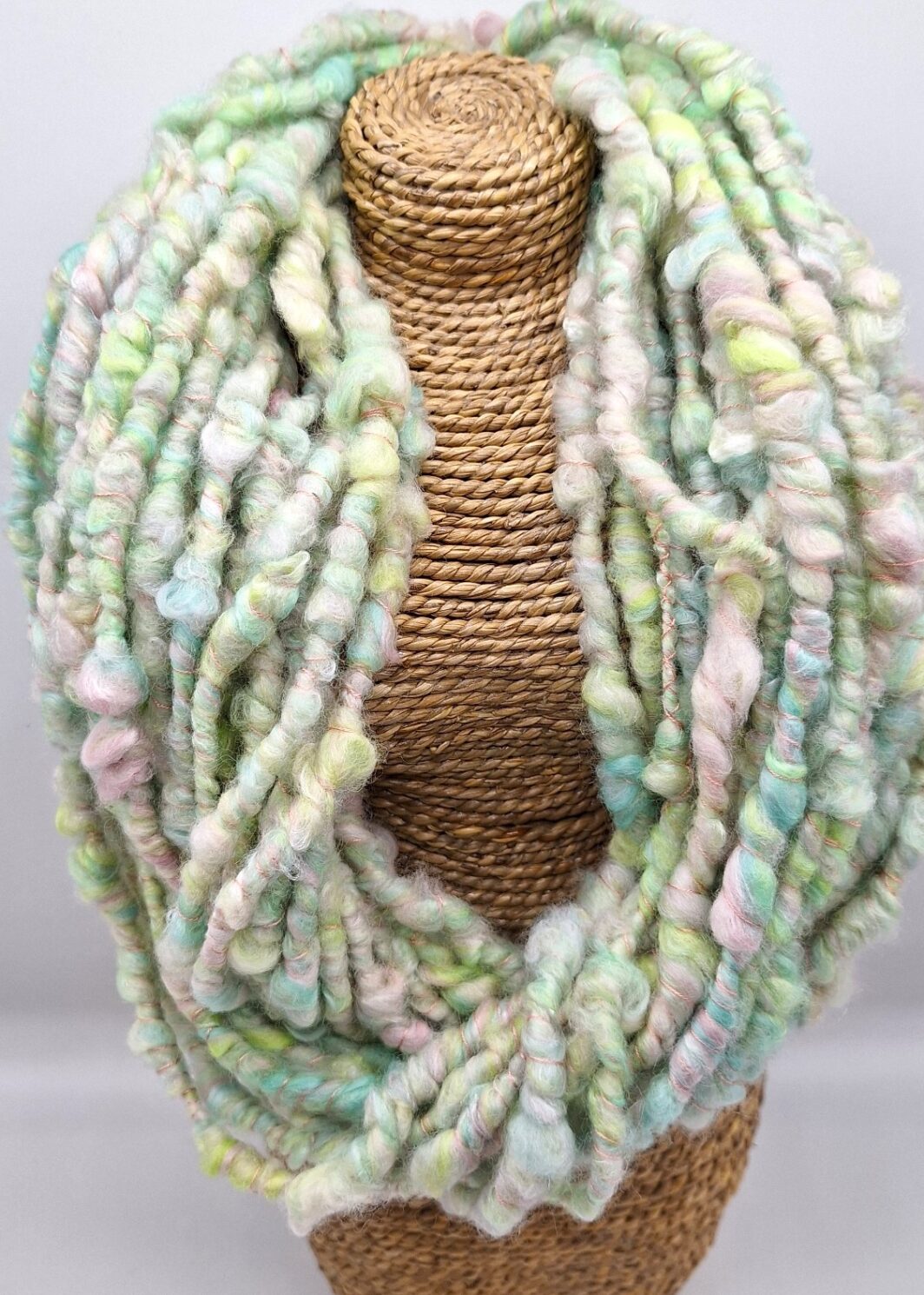 Hand spun merino and silk cowl neckwarmer in pastel shades of green, pink, blue and white on a rattan bust.