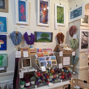 An area inside a shop with felt art and hand spun cowls in various colours hanging on the wall below which is a display of colourful carded batts, merino fibre and hand spun yarn. Between the two is a display of greeting cards.