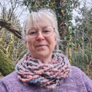 Middle aged white woman in glasses standing among trees wearing Araminta Greaves Fibreista hand spun merino wool cowl neck warmer in shades of peach, bluey grey and deep pink.