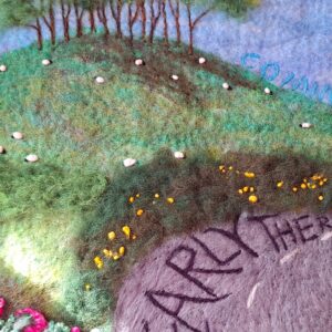 Detail of a felt picture of Cookworthy Knapp by Araminta Greaves Fibreista. Felt picture depicts trees on a hill with sheep grazing in the fields and lettering spelling out Nearly There on the road and Coming Home in the sky.